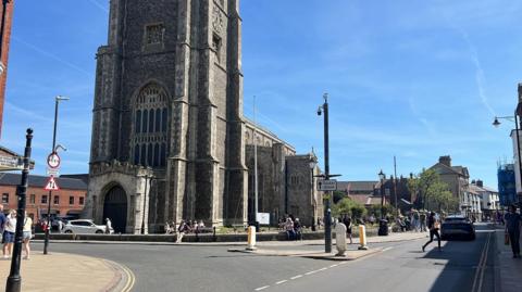 Part of Church Street in Cromer. There are people walking along the pavements and a car is driving along the road. There is a large church in the image.