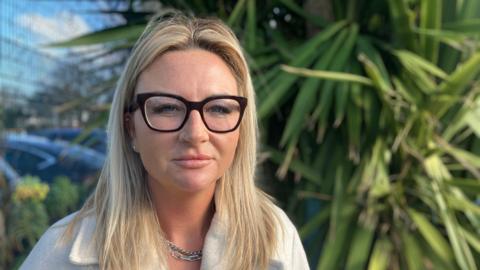 Charlotte Smart stood in front of a tree. She is wearing a white top and black glasses, and looking directly at the camera. 