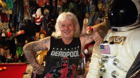 Man with long grey hair in a Deadpool vest leaning on a Nasa statue
