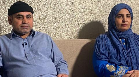 Israr Hussain, on the left, and his wife, Tasleem Akhtar, are both wearing blue. They are sitting on a sofa by a wall.