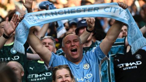 Manchester City fan wearing a blue team shirt holds a scarf with the club's name on it over his head in a crowd