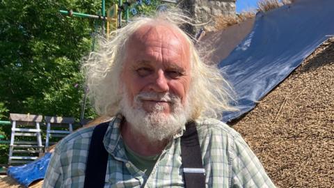Alan Jones wearing a checked green and white shirt with black braces. He is standing in front of a thatched roof. 