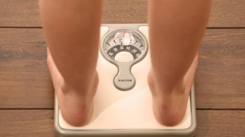 Person standing on a set of weighing scales. The backs of the legs and feet are visible on the scales. The scales are white and have been placed on a wooden floor.