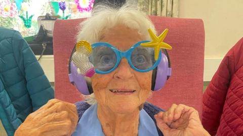 Olive Parsons wears a pair of blue novelty sunglasses that have seashells in the frame. She seated on a pink chair and is smiling while holding her hands up in celebration