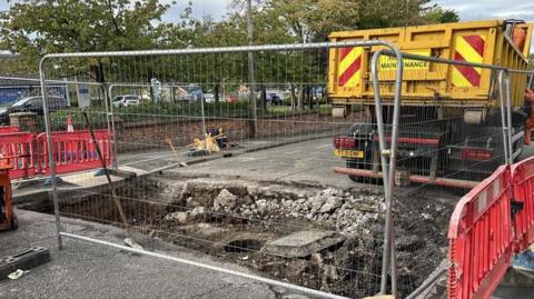 A large section of road has been dug up and metal fencing has been put on the road to prevent drivers getting too close. A yellow Highway Maintenance truck is parked nearby.