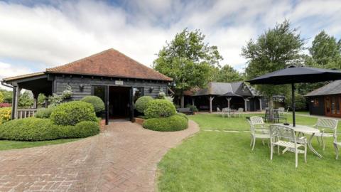 The Old Essex Barn near Kelvedon. A barn building with dark panel weatherboards on the outside, with a slate roof, is on the left of the image, with several tables and chairs on an area of grass to the right.