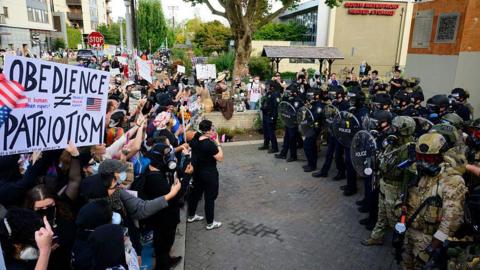 Protesters stand with signs - some wearing masks - stand in a line facing off with authorities in military fatigues and police uniforms, some carrying rio shields, in Portland, Oregon. 