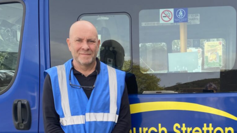 A man with a bald head and blue reflective jacket in front of the side of a blue minibus with black windows and yellow writing
