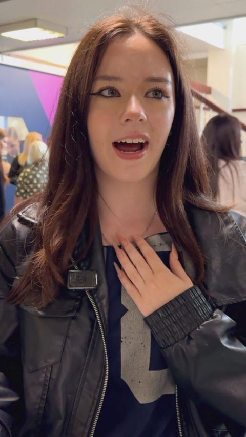 A girl who has received her GCSE results today is standing overjoyed. She has dark red hair, and is wearing a black zipped leather jacket.