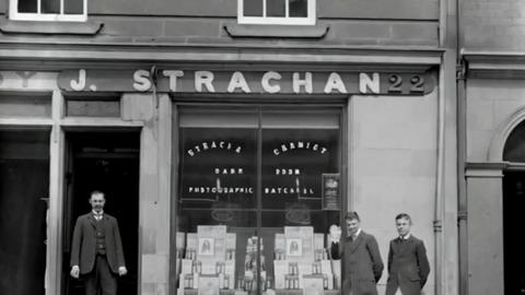 Three men in suits outside a shop called J.Strachan