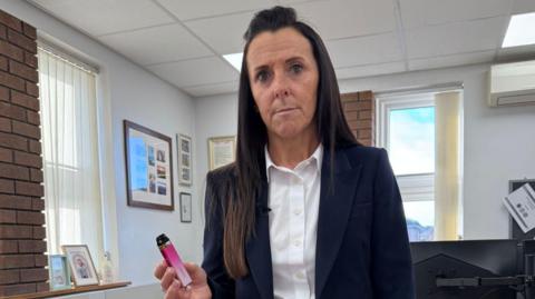 Head teacher wearing navy suit holds up a pink vape