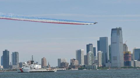 Aircraft trail red, white and blue smoke above waterfront skyline, passing US Coast Guard vessel