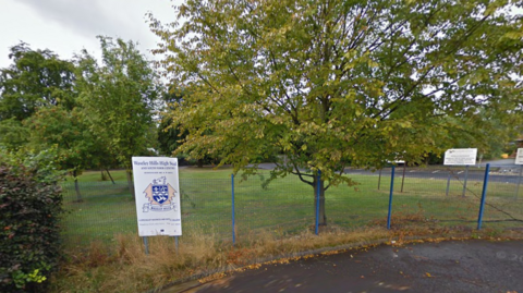 A photo of a school sign, white with a crest on it and reading Waseley Hills High School, on a blue security fence with trees on grass behind. 
