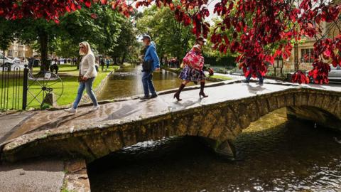 A stone bridge crossing the River Windrush with grass on one side a green and red leaved trees. People are crossing the bridge on a sunny day.
