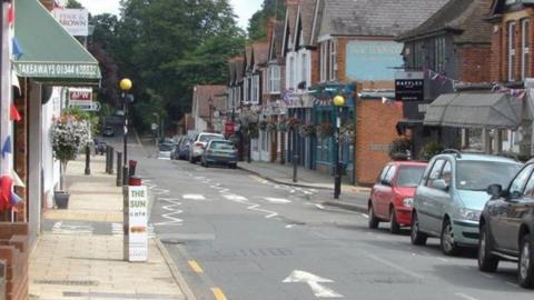 High street with road running through - parked cars to the right and a zebra crossing the centre shops/buildings run along both sides.