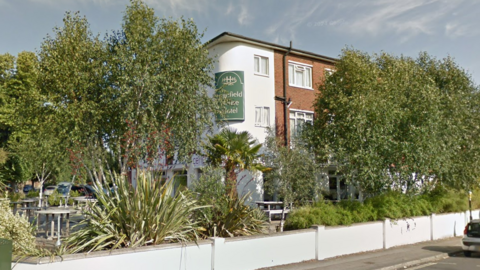 Street View image of Highfield House Hotel, a three storey building of brick and white render set back from the street corner behind a low white wall. The garden is filled with trees, palms and tropical-looking plants.