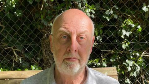 Steve Clare has short grey hair and is looking at the camera. He has a short white beard and he is sitting outside on a bench. There is a hedge behind him through a fence.