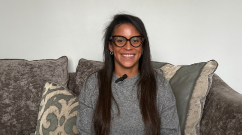 The image shows a woman sat on a couch, smiling at the camera. There is a microphone clipped to the collar of her jumper and she is wearing glasses with black frames. There are cushions behind her and the wall of the room is white.