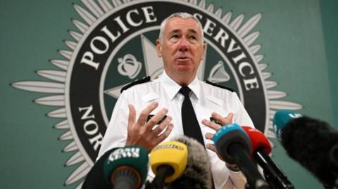 PSNI chief constable Jon Boutcher, in white shirt and black tie, in front of PSNI crest, with microphones ahead of him