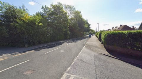 The junction of two roads. There are hedges either side of the road with a few rooftops visible in the distance.