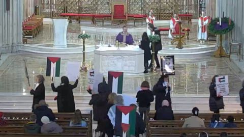 Protesters holding up placards with the Palestinian flag on them standing at the front of a cathedral.  A priest in purple robs is standing behind an alter behind them.