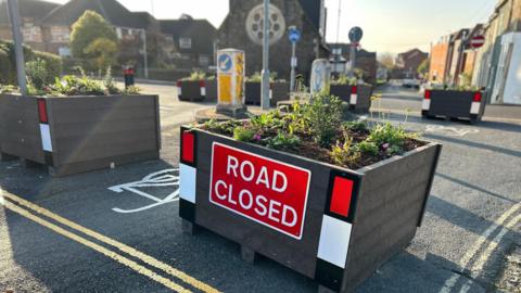 Several planters on a street with a red "road closed" sign illuminated,