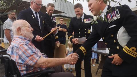 An elderly man in a check shirt sitting in a wheelchair and shaking hands with a man in a naval uniform wearing a number of medals.  In the background there are a number of other men in naval uniforms along with civilians and children
