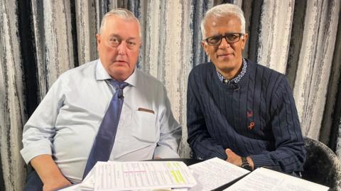 Two men sat together with papers arranged on a desk  