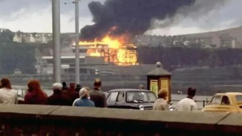 An old photograph of people looking on at the Summerland Complex, which is ablaze with a thick plume of black smoke siring from it.