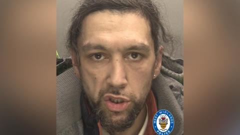 Luke Dhear - a custody image of a man with long brown hair with grey streaks that is tied back and a brown and grey short beard, he is wearing what appears o be a grey coat with a black and orange top underneath. He has a stud earring in his left ear and 