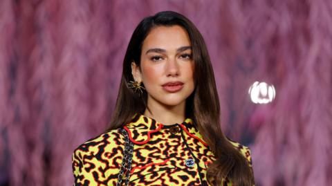 Dua Lipa poses with her dark brown hair straightened and out, while wearing a yellow, black and red Chanel jacket and a gold spiky earring.