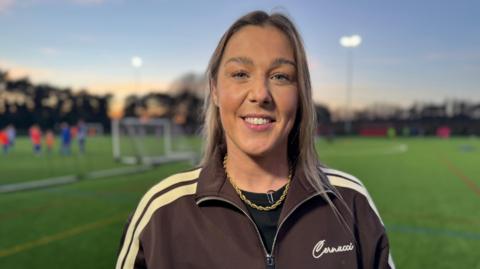 Mary Earps, wearing a fashion tracksuit top, looking at the camera while stood in front of an all weather football pitch