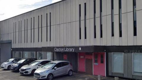 Clacton Library. The building has a red door and several cars parked outside it. The building grey. It has a sign that says Clacton Library at the entrance.