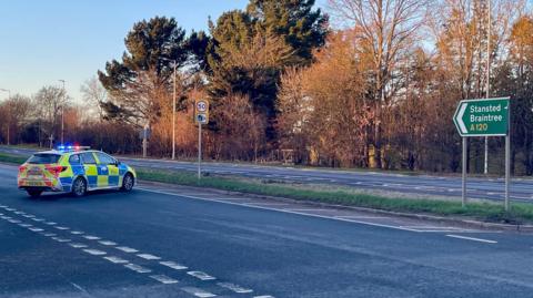 A police car is sit across a road with its lights on. There is a junction on the left of the image and a sign on the right which says Stansted and Braintree on it.