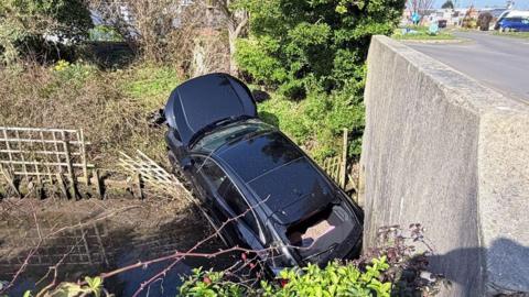 A black car is in a ditch in water with its front on the bank and its bonnet open. It is near a bridge. A fence is broken and the ditch has trees and undergrowth on its banks.