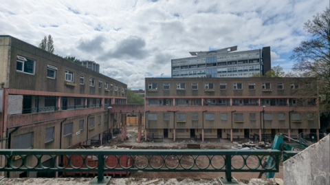 A shot looking down onto a building site. Some rather dilapidated blocks of flats run along the edges. There appears to be a large hole in the middle of the site and a blue-green crane and what appears to be a large red skip are nearby.

