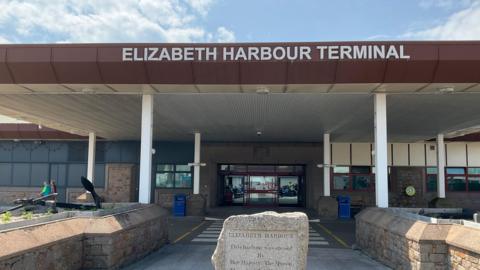 A picture of the front of Elizabeth harbour terminal. It is a large building with a glass entrance and a stone in front of it.