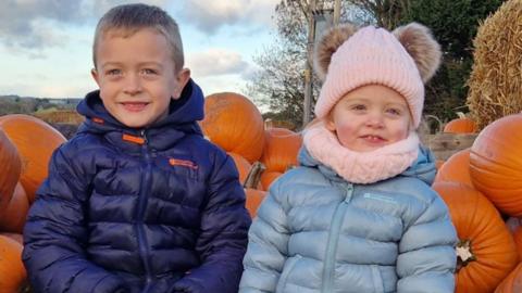 Ted and Hollie sit in a pumpkin patch surrounded by pumpkins. Ted wears a dark blue puffer jacket, he has short blonde hair. Hollie wears a light blue puffer jacket, a pink bobble hat and a matching pink scarf. They both smile at the camera