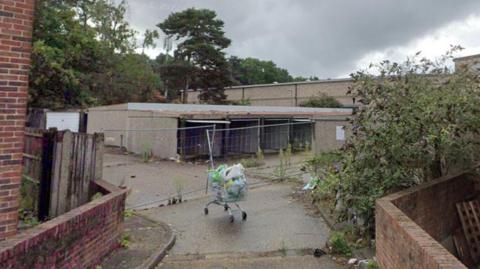 A set of derelict garages which are falling apart. A road leads up to the garages, and an abandoned shopping trolley sits in the middle of the road.