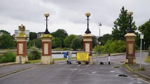 Several pillars signify a road entrance to a park. There is a yellow sign in front of one of the pillars that reads: "Poole Park closed to through traffic." A lake and trees can be seen in the background. 