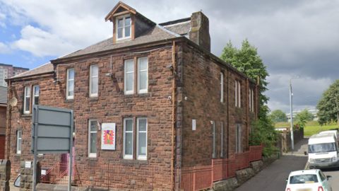 A red brick building, with railings around it. It is the last building on the street, with a road running upwards next to it and cars parked beside it.