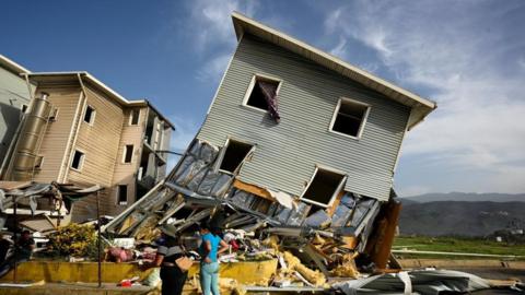 A building that is wonky and fallen and now half destroyed with a huge pile of rubble in front of it 