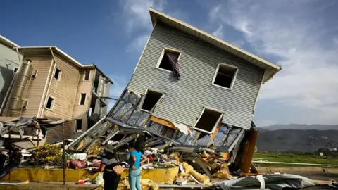 A building that is wonky and fallen and now half destroyed with a huge pile of rubble in front of it 