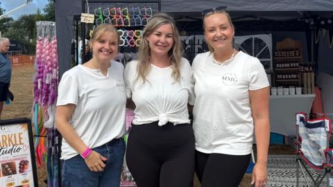 The image shows three women wearing white t-shirts standing outside a stall at the West Show.