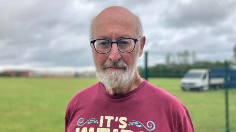 Tony Forster looking at the camera with a glum expression. He is wearing glasses and a dark red T-shirt. Behind him is the partially built green metal fence and a van on a field. 