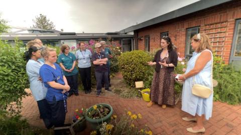 Sobell House Hospice staff gathered around in their inpatient unit garden as part of the time capsule ceremony. There are many flowers around. It is an overcast day.