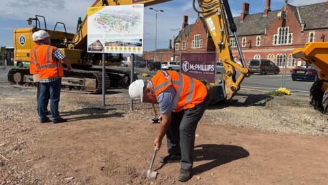 Councillor Philip Price wears a white helmet and orange hi-vis vest and uses a spade to dig on the site during the ground-breaking ceremony. Two other men are standing to one side and there is a digger behind him, with a map of the site on a board.