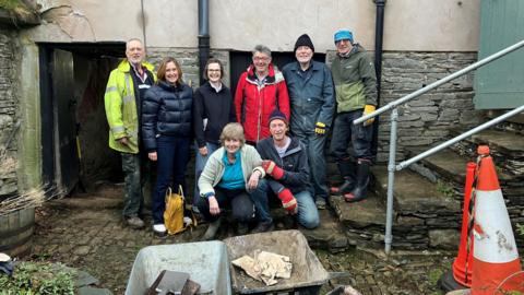 Eight people are posing in front of Yew Tree Hall. They are a little muddy after working on the redevelopment.