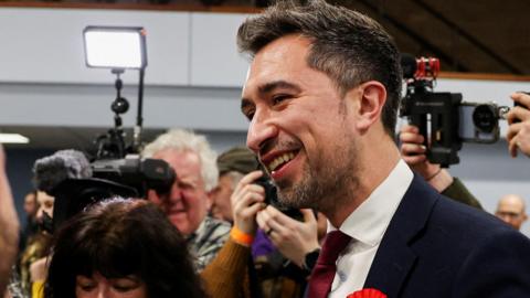 Damien Egan at an election count. He is wearing a red rosette and is smiling. He is surrounded by people.