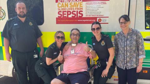 Roger Field sitting in front of an ambulance. The paramedics who helped save his life, Simon Sayers, Steph Oldham and Alicia Ray, are standing around him, as is his wife Lynn.